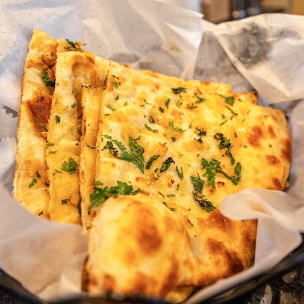 Close-up of golden Indian naan bread slices, garnished with fresh green herbs, served in a basket.