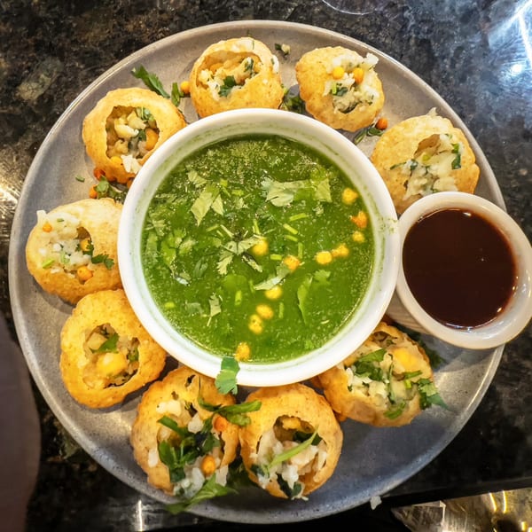 A plate of Indian pani puri, with a bowl of green mint water and a small bowl of tamarind chutney.