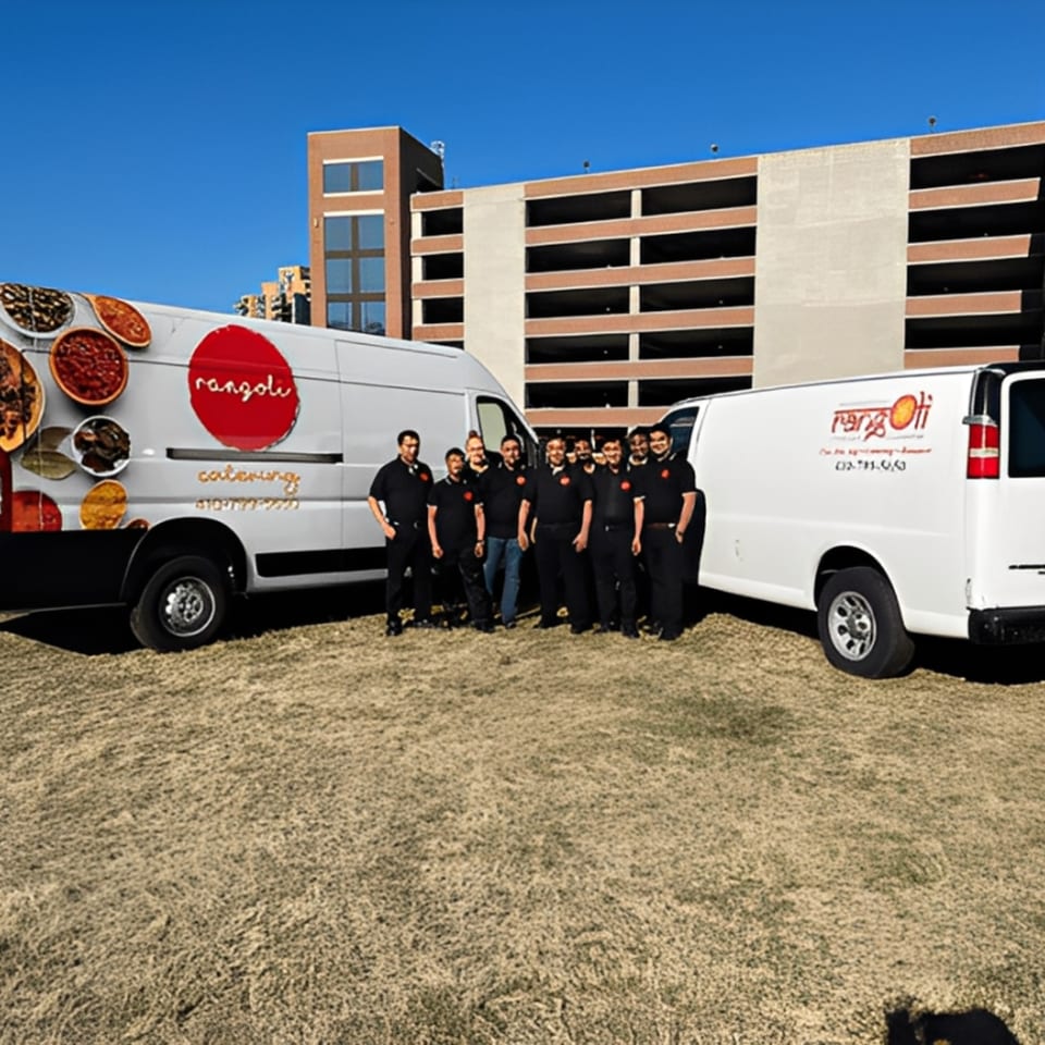 Rangoli Restaurant catering team stands proudly between two branded catering vans on a sunny day.