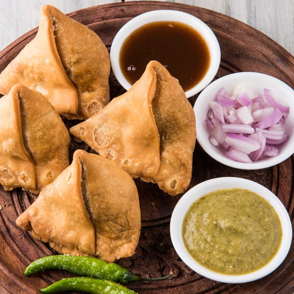 Golden-fried samosas, green chili peppers, and bowls of chutney and chopped red onions on a wooden tray.