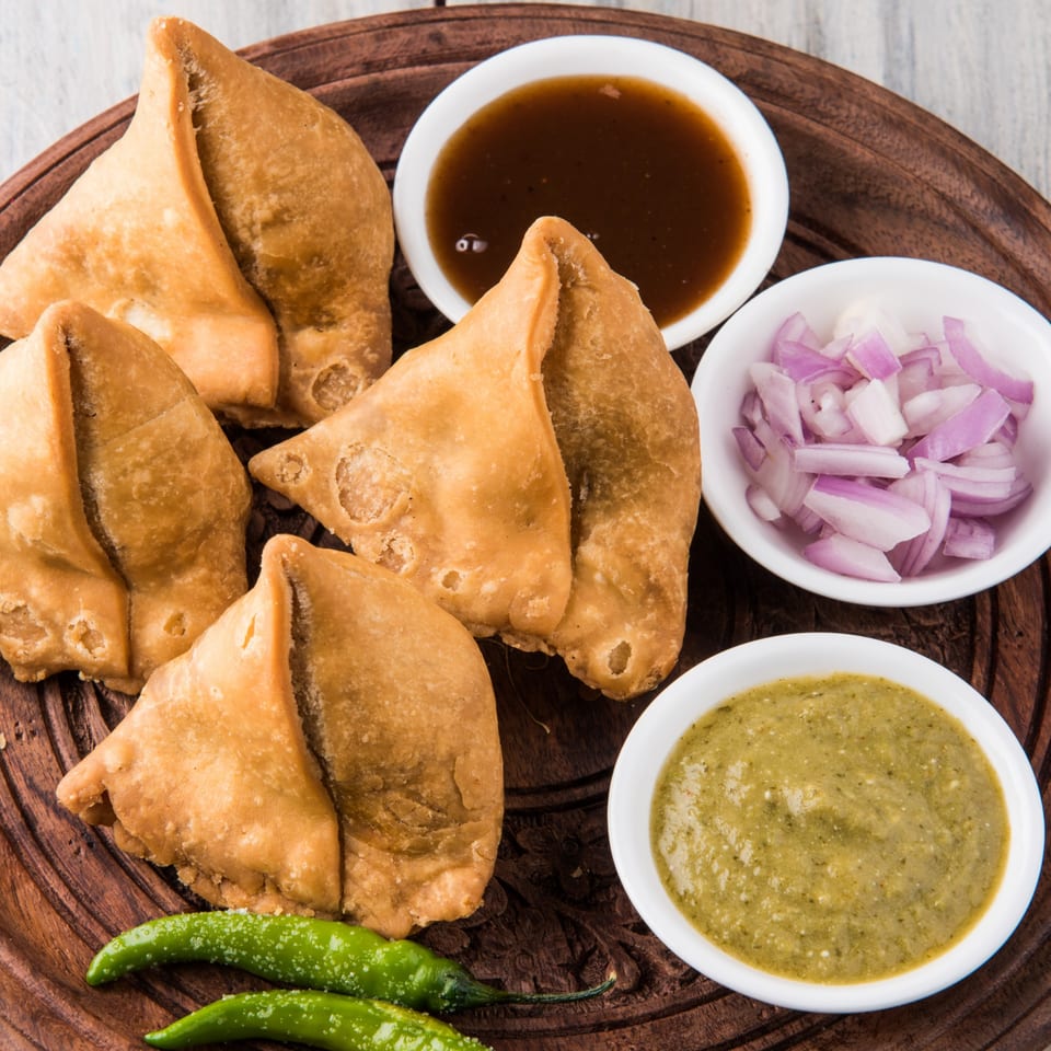 Golden-fried samosas, green chili peppers, and bowls of chutney and chopped red onions on a wooden tray.