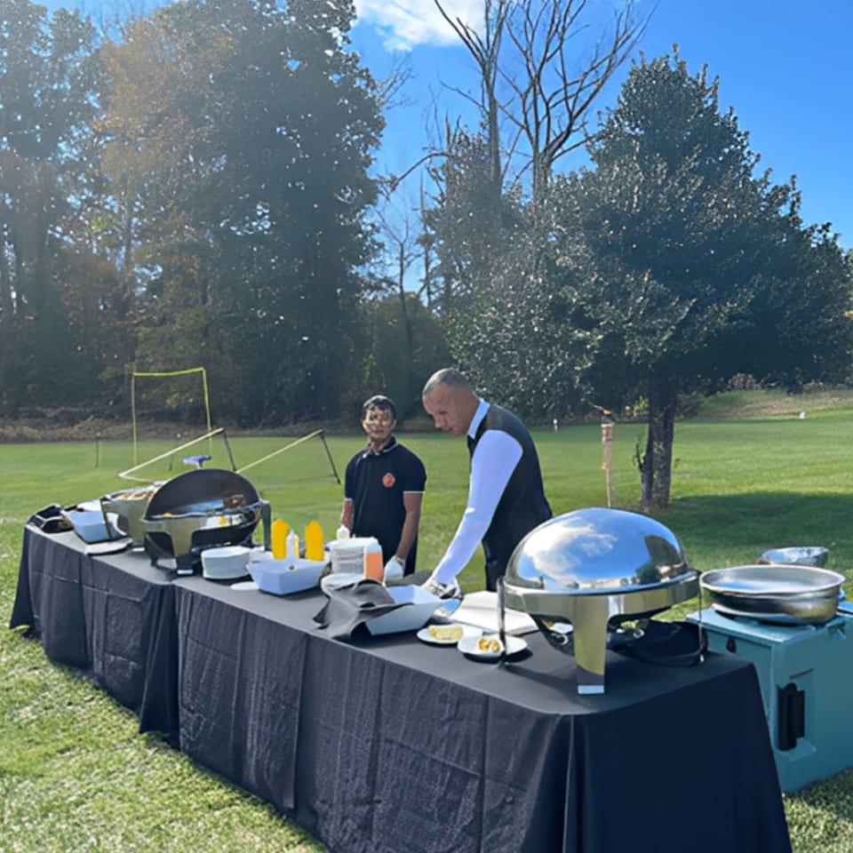 Two Rangoli staff members serving Indian food from a buffet line outdoors on a sunny day.
