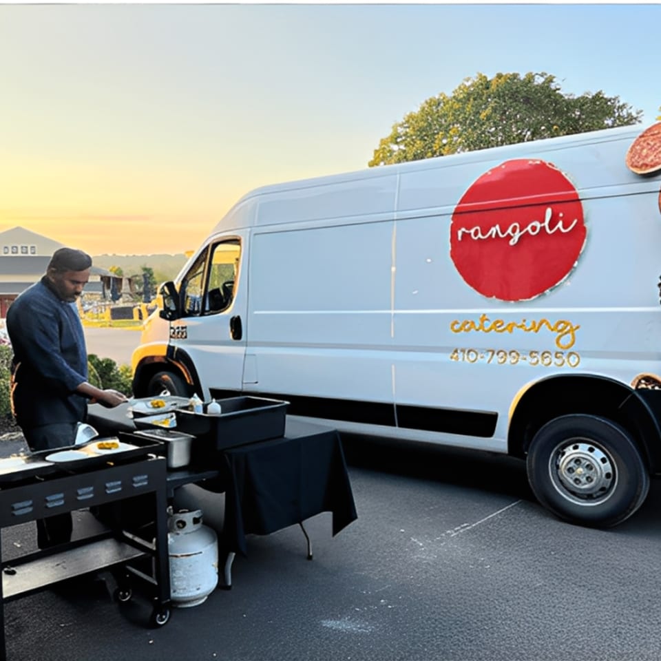 A chef prepares food on a grill next to the Rangoli catering van at sunset.